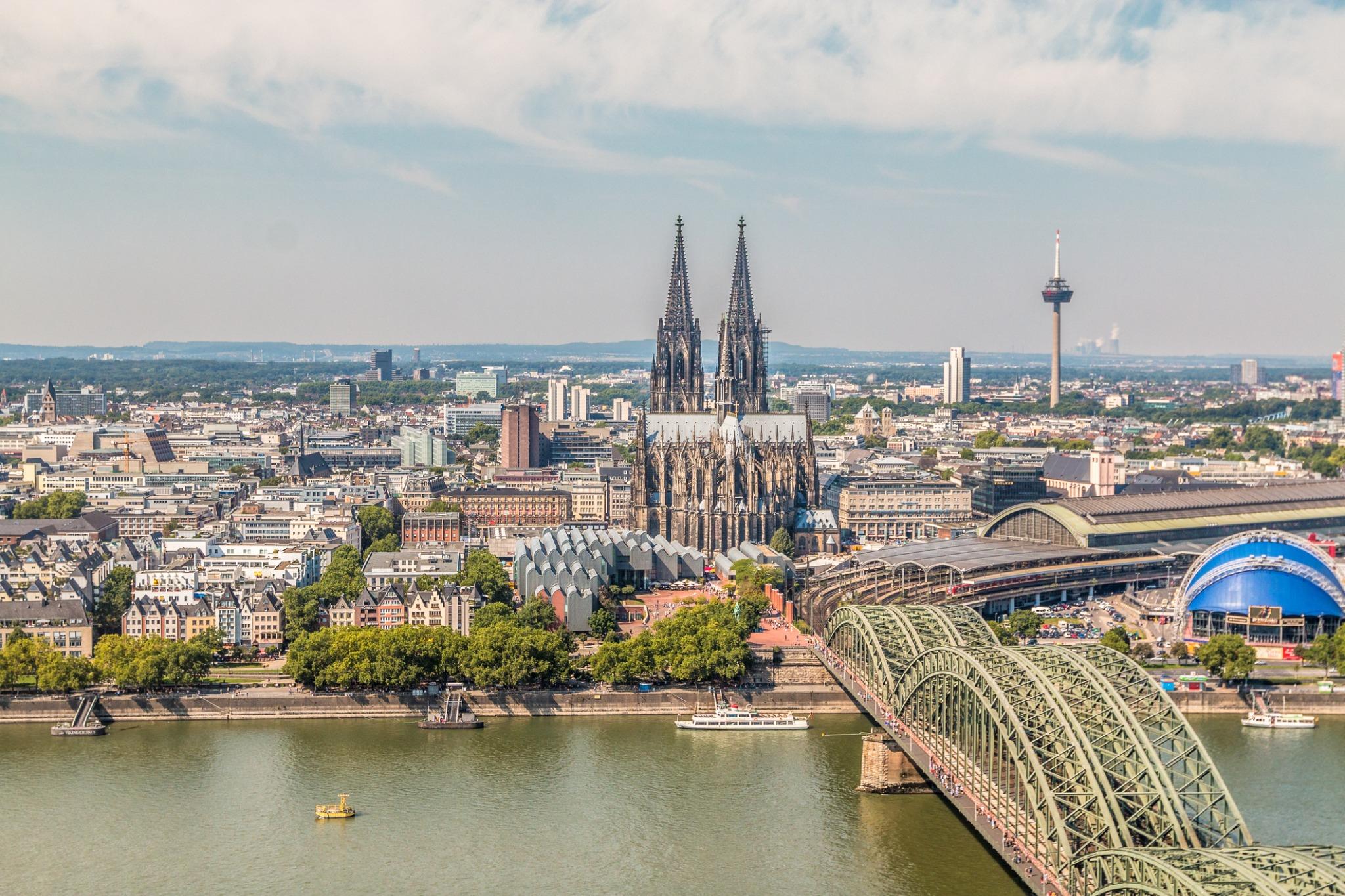 Aerial view of Cologne, Germany, featuring the Cologne Cathedral, Hohenzollern Bridge, and surrounding cityscape along the Rhine River.